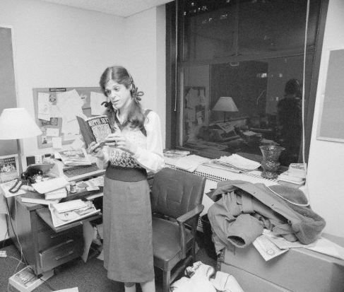 Gilda Radner stands in a messy office reading a book